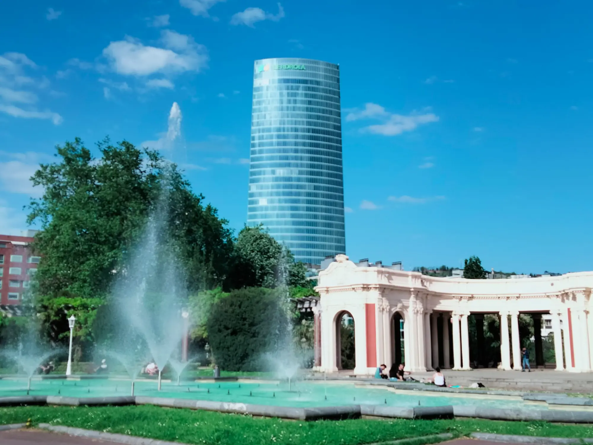 Vista de un parque urbano con una fuente en primer plano, un templete clásico y la torre Iberdrola al fondo bajo un cielo azul.